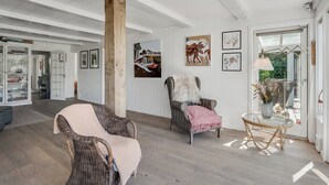 Une pièce spacieuse avec un plafond haut, des poutres en bois, une grande fenêtre et un coin salon confortable avec un fauteuil et une table.