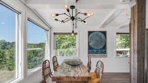 Un coin repas avec une table en bois, des chaises en osier, un lustre et de grandes fenêtres donnant sur un paysage verdoyant.