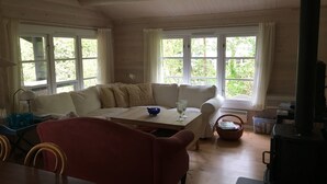 A cozy room with a wooden ceiling, a sofa, a red chair, a table, and a fireplace. - Log house near a child-friendly sandy beach (Nykøbing Sjælland)