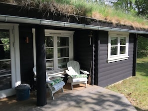 A cabin with a porch, two white chairs, a trash can, and a window.