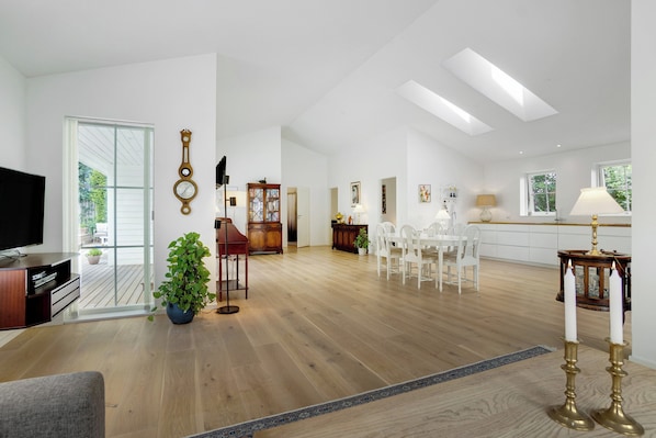 A spacious living area with a wooden floor, a television, a dining table with chairs, and a large window.