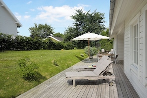 A wooden deck with a lounge chair and an umbrella, a small garden, and a white building.