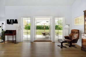 A room with a wooden floor, a brown leather chair, a desk, a lamp, and a view through glass doors to an outdoor patio with a table and umbrella. - New and cozy house by the beach and forest (Hornbæk)