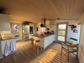Une cuisine avec un plafond en bois, des appareils en acier inoxydable, un Ăźlot central avec des tabourets et une vue sur lâextĂ©rieur, visible Ă travers la porte.