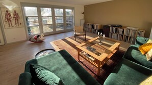 Uma sala de estar ampla com uma grande porta de vidro deslizante, mesa de centro de madeira, sofás verdes e uma estante cheia de livros e discos.