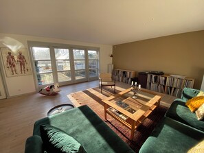 Un salon spacieux dotĂ© dâune grande porte-fenĂȘtre coulissante, dâune table basse en bois, de canapĂ©s verts et dâune bibliothĂšque remplie de livres et de disques.