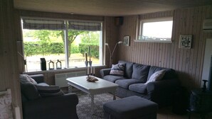 Una sala de estar con un sofá, un reposapiés y una mesa. Hay ventanas con cortinas, un radiador y un marco de cuadros en la pared.