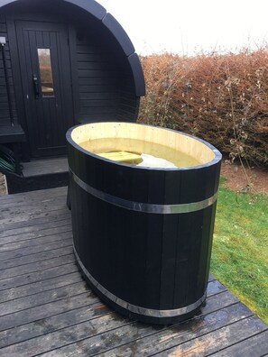 Ein großes, schwarzes Holz-Whirlpool auf einer Holzdiele mit einem schwarzen Gebäude im Hintergrund.