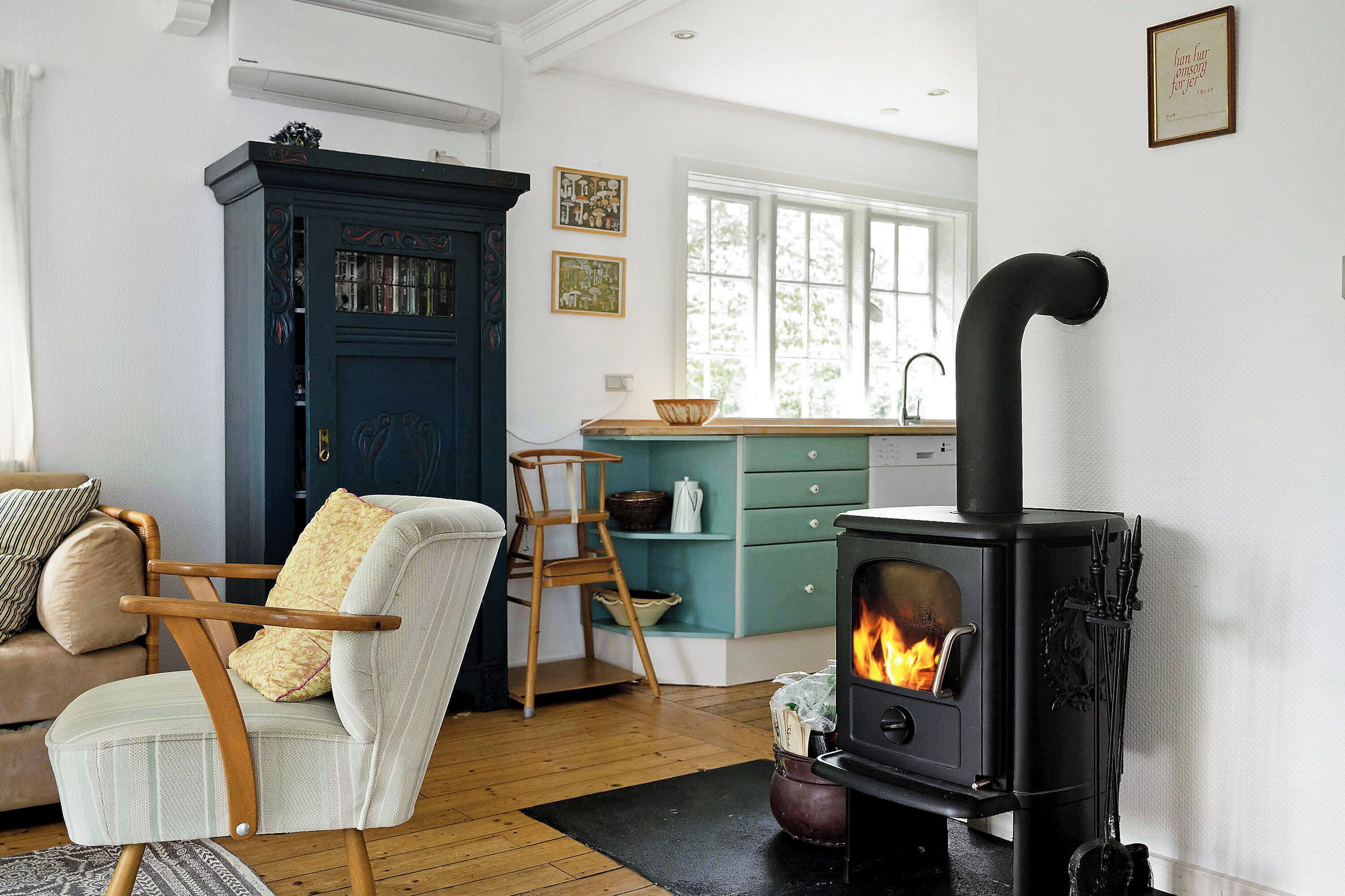 A cozy living room with a sofa, a chair, a wooden floor, a black stove with a chimney, and a window.