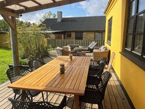 Una terraza de madera con una mesa y sillas, un espacio cubierto con una pérgola, y una casa al fondo.
