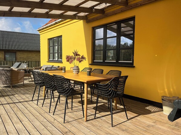 Une terrasse en bois avec une table et des chaises, un mur jaune et une fenêtre.