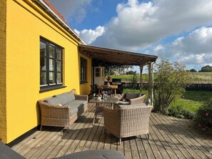 Un bâtiment jaune doté d’une terrasse en bois, de meubles en osier et d’un espace barbecue.