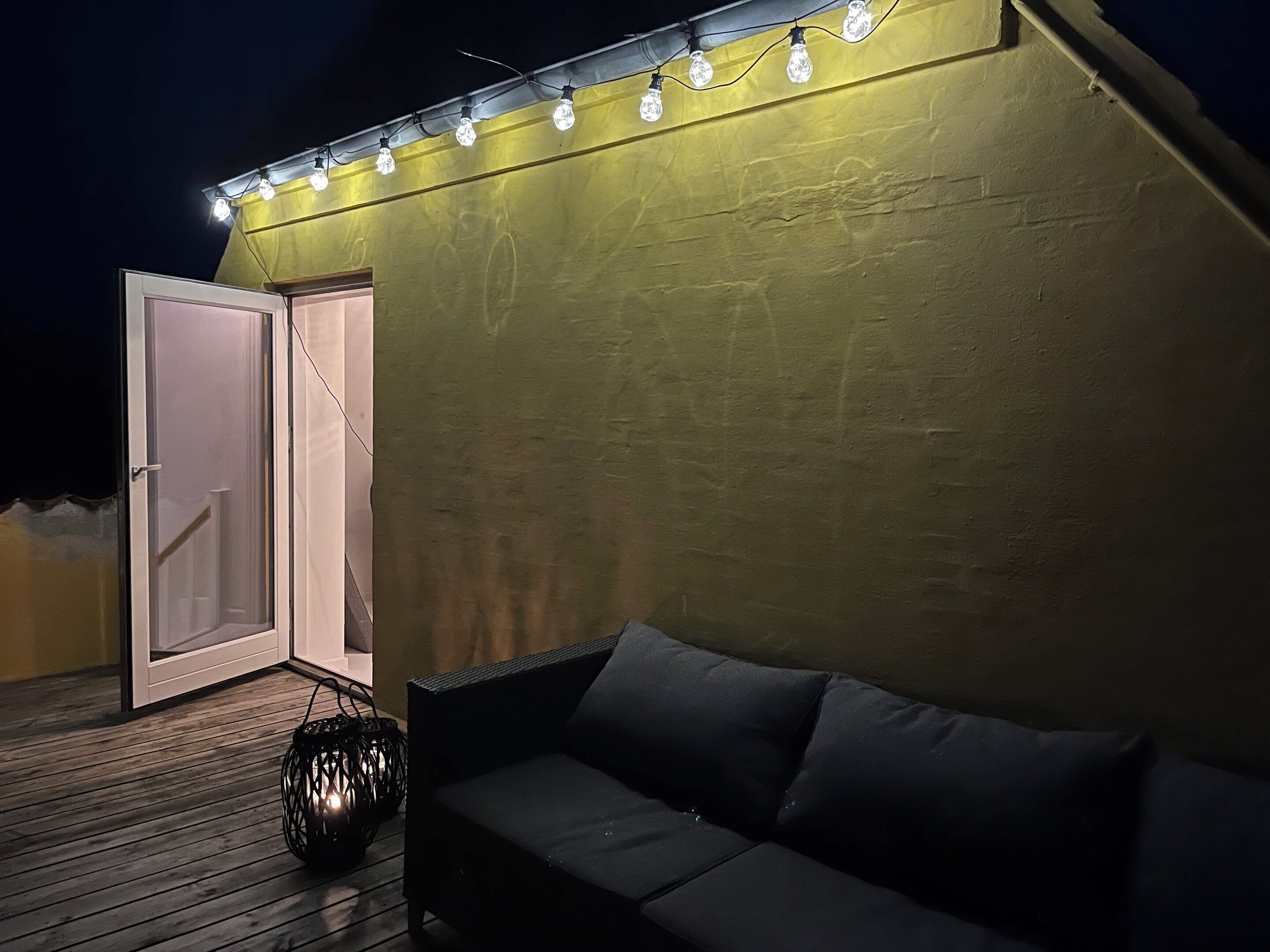 A cozy outdoor seating area with a sofa and a lantern, a door leading to a covered space, and string lights on the wall.
