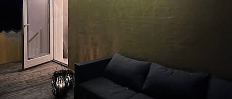 A cozy outdoor seating area with a sofa and a lantern, a door leading to a covered space, and string lights on the wall.