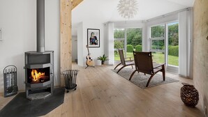 A room with a wood-burning stove, a guitar, a chair, and a view of greenery through large windows.