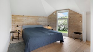 A bedroom with a wooden headboard, a bed with a blue bedspread, a nightstand with a lamp, a window with a view of greenery, and a small stool.