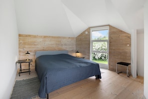 Un dormitorio con cabecera de madera, cama con colcha azul, mesita de noche con lámpara, ventana con vista a follaje y un taburete pequeño.