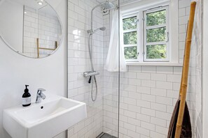 Ein Badezimmer mit weißem Waschbecken, einem Spiegel, einer Duschkabine mit Glastür und einem Fenster mit Blick auf Bäume.