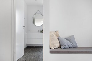 Un baño moderno con tocador blanco, un espejo redondo y un banco con almohadones.