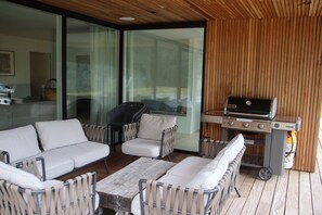 An outdoor seating area with a wooden deck, a white sofa, two armchairs, and a table. There is a barbecue grill with a gas tank and a control panel. - Holiday home on Bornholm close to sea and beach (Aakirkeby)