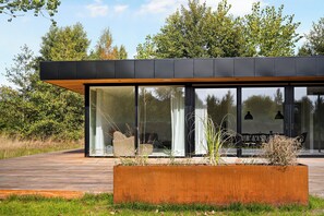 Un bâtiment moderne doté de grandes fenêtres en verre, d’une terrasse en bois et d’un jardinier avec des plantes.
