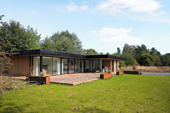 Une maison moderne dotée de grandes fenêtres en verre, d’une terrasse en bois et d’une cour gazonnée.