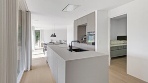 A modern kitchen with a central island, a dining area with a table and chairs, and a bedroom with a bed and nightstand. - Holiday home on Bornholm close to sea and beach (Aakirkeby)