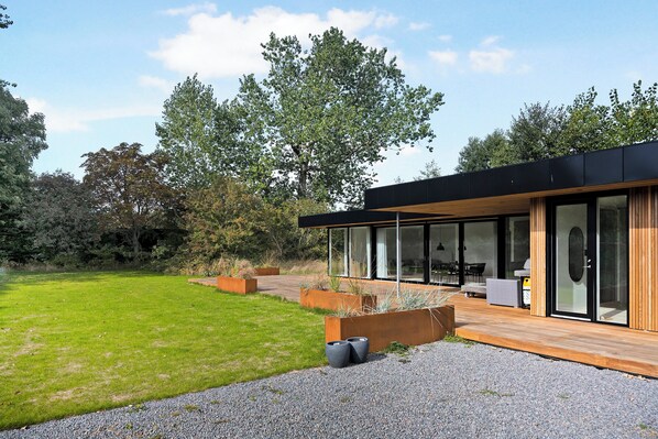 Une maison moderne dotée d’une terrasse en bois, de grandes fenêtres et d’une cour gazonnée.