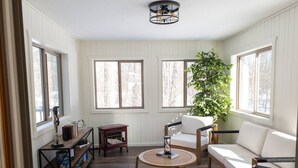 A room with a wooden floor, white walls, a round table, a sofa, a bookshelf, and a large window with a view of trees.
