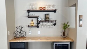 A compact kitchen with a wooden countertop, a black cabinet, a mini fridge, and a wall-mounted shelf with glasses and a whiskey bottle.