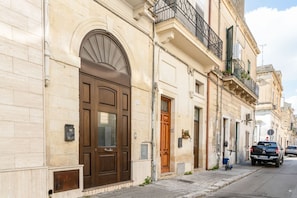 A street view of a building with a large arched entrance, a balcony with a black railing, and a parked car on the adjacent street.
