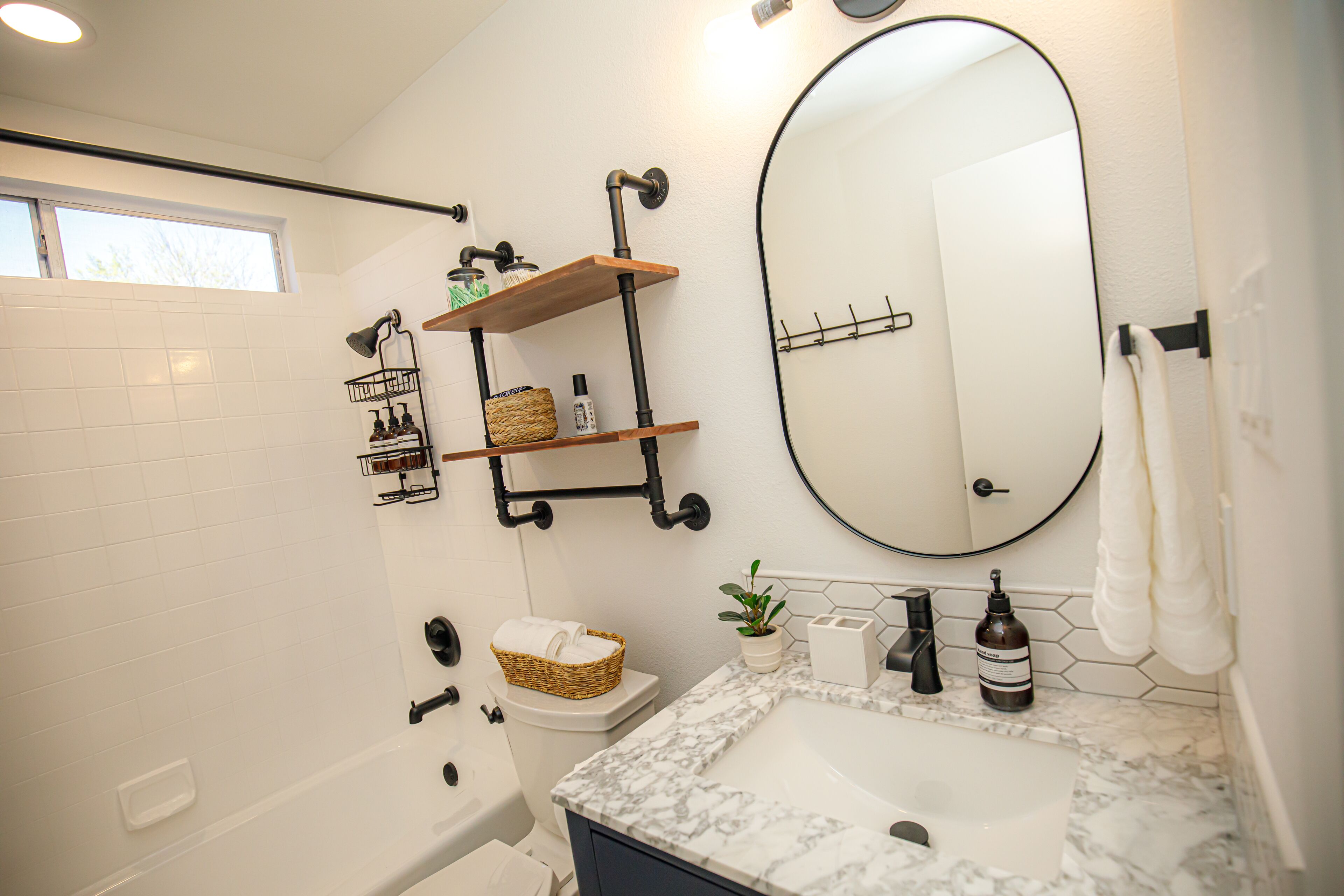 A modern bathroom with a white tiled wall, a large oval mirror, a marble sink, and a built-in toilet. There are shelves with toiletries and a basket on the toilet.