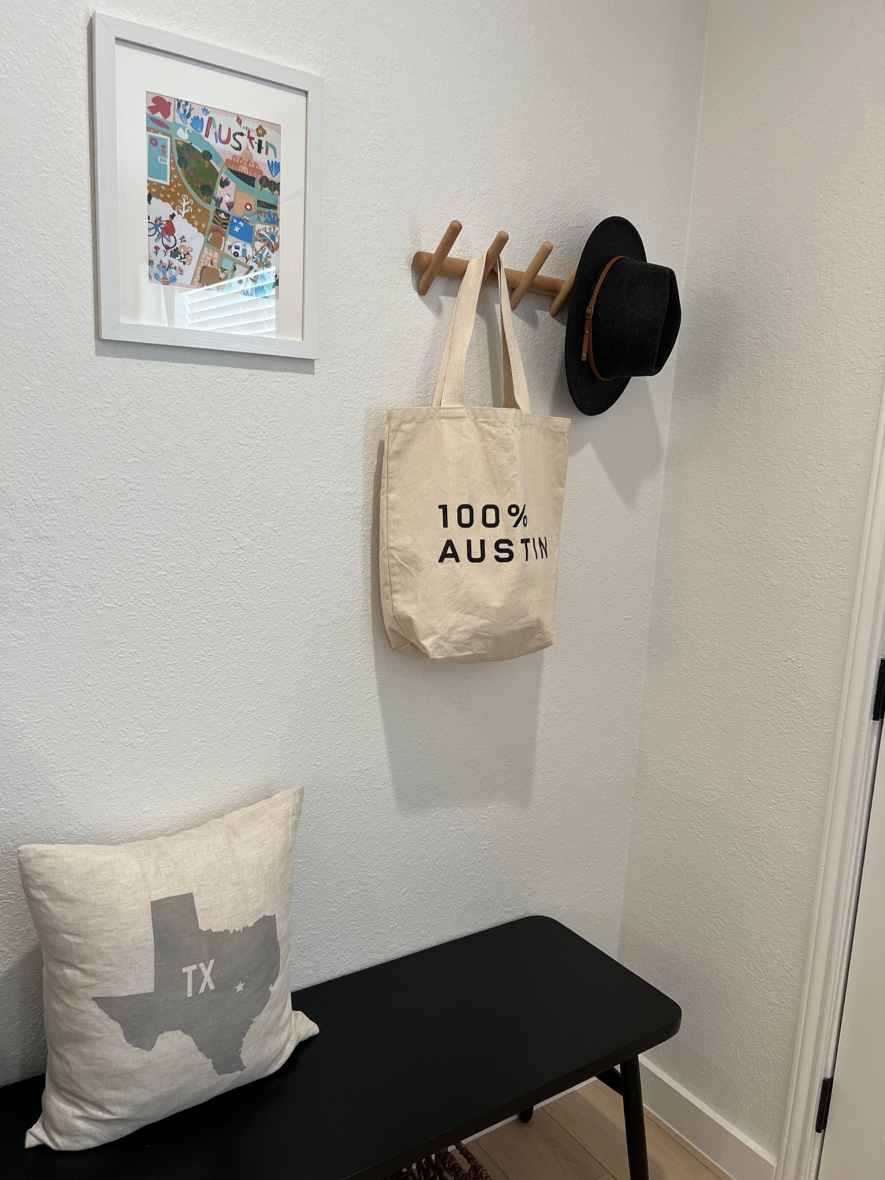 Una habitación con un banco negro, un perchero de pared, un mapa enmarcado, una bolsa de tela y un cojín con el contorno de Texas.