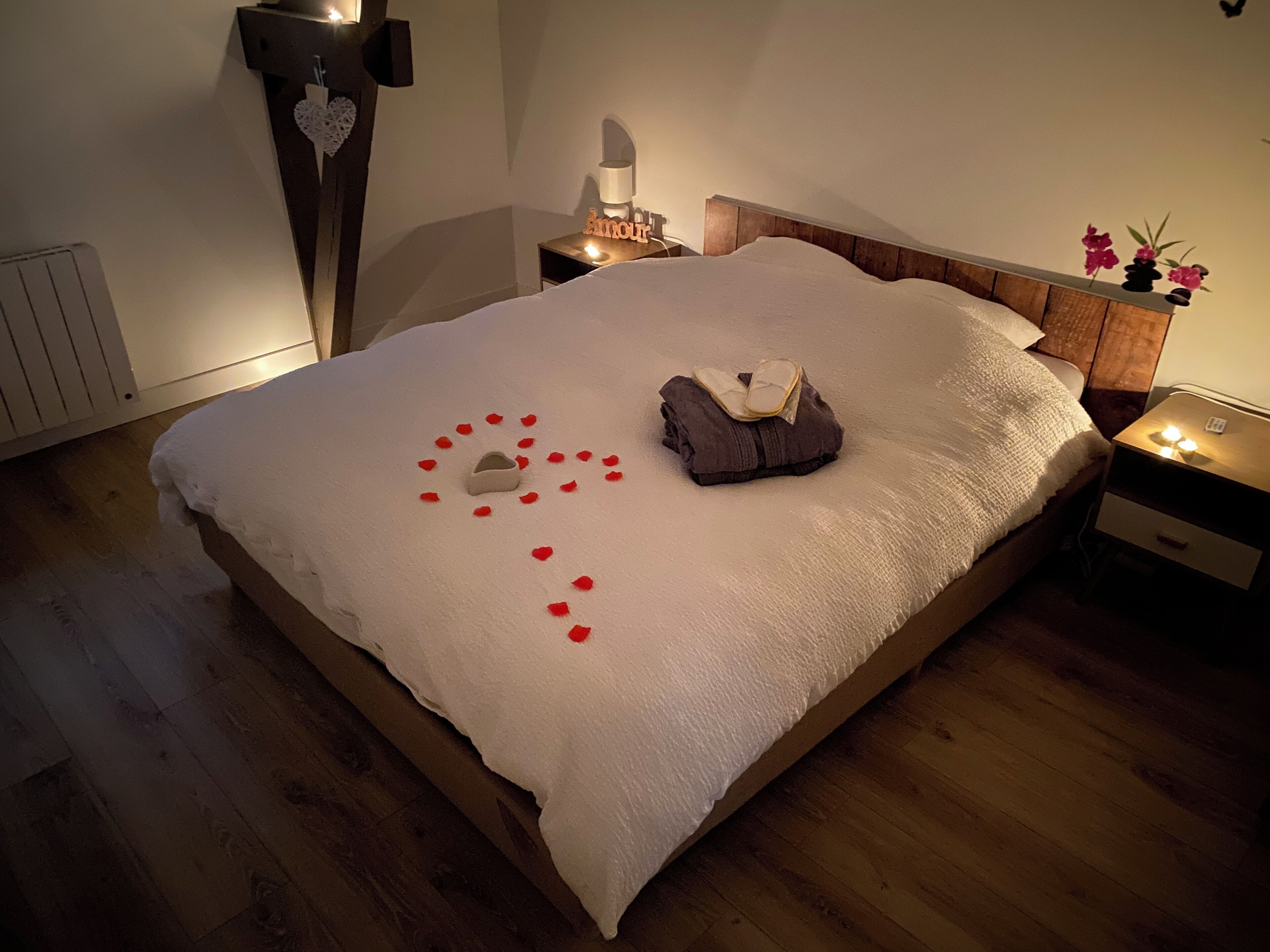 A neatly made bed with a heart-shaped arrangement of rose petals and a small heart-shaped object on the bed.
