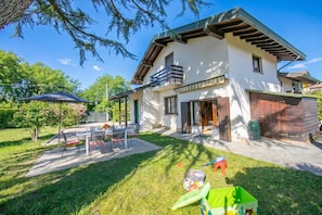 Ein zweistöckiges Haus mit Balkon, einer Terrasse und Gartenmöbeln sowie einem grünen Spielbereich.