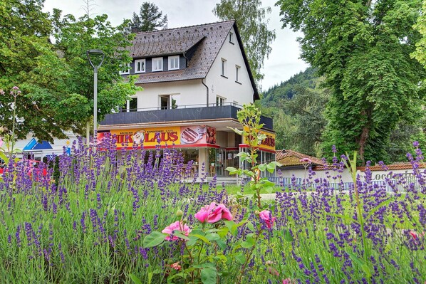 A two-story building with a balcony, surrounded by lush greenery and purple flowers.