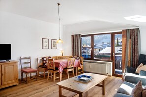 Una sala de estar con una mesa de madera, sillas, un sofá, un televisor y una ventana con vistas a edificios y montañas.