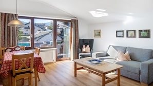 A living room with a sofa, a table, a chair, and a window with a view of houses and mountains. - Holiday Apartment "Haus Excelsior Top 40" with Mountain View, Balcony & Wi-Fi (Seefeld in Tirol)