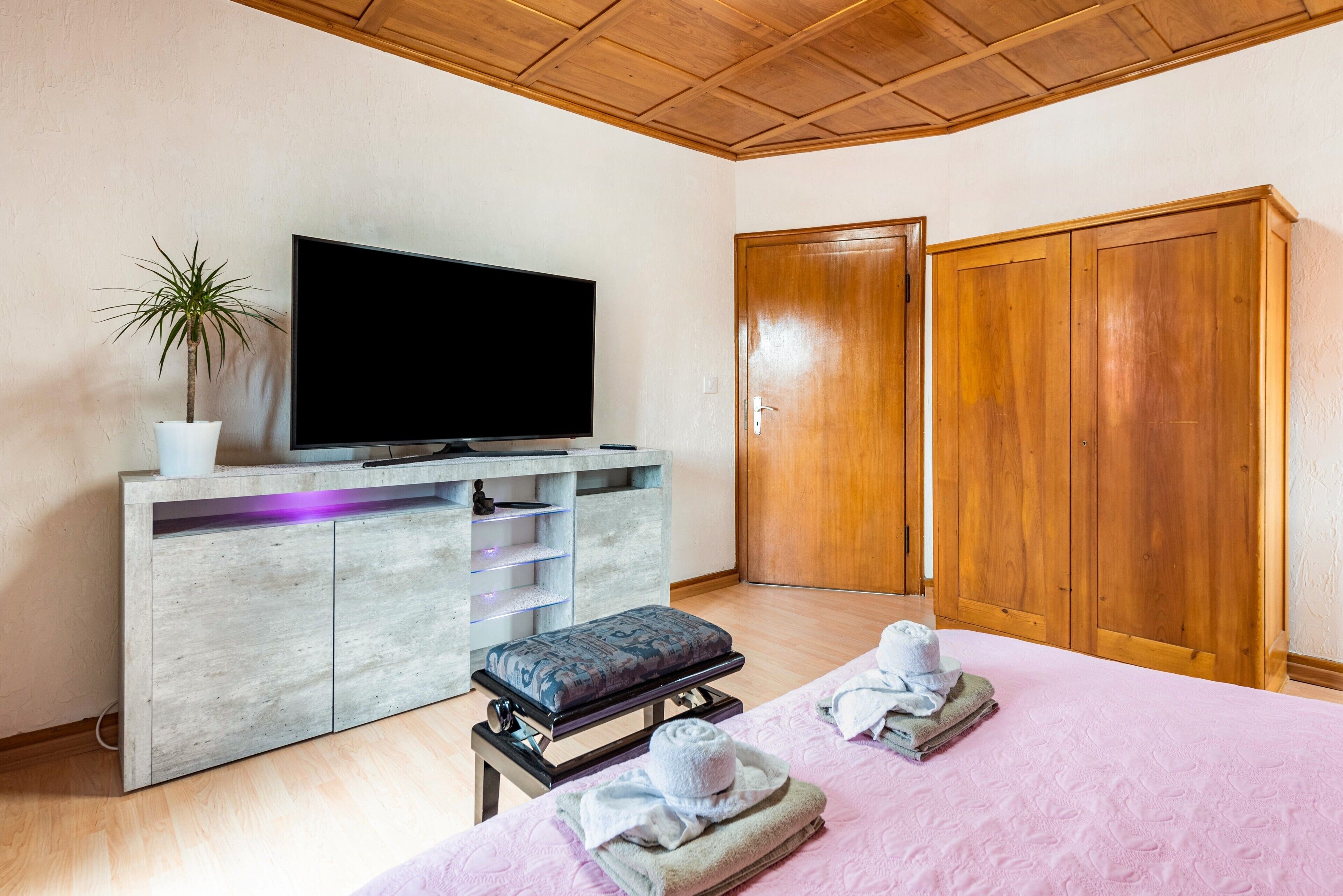 A bedroom with a television, a wooden wardrobe, a bench, a potted plant, and towels on the bed.