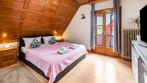 Un dormitorio con techo inclinado, una cama con colcha rosa, dos mesitas de noche con lámparas, un radiador y vistas a un edificio rojo a través de una puerta de cristal.