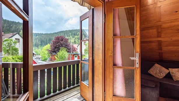 Puerta de madera que da a un balcón con vistas a una colina boscosa y casas.