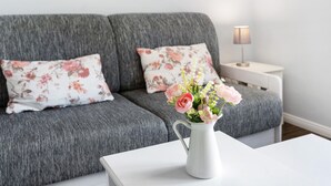 Een grijze bank met bloemkussens, een witte bijzettafel met een lamp, en een witte vaas met bloemen op een witte tafel.