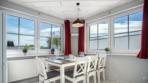 Un espace repas comprenant une table et des chaises, de grandes fenêtres avec des rideaux et une vue sur l’extérieur.
