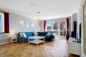 Un salon avec un canapé bleu, une table basse blanche, un téléviseur et une fenêtre ornée de rideaux rouges.