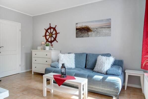 A living room with a blue sofa, white drawers, a white table with a red cloth, a ship's wheel, and a beach painting.