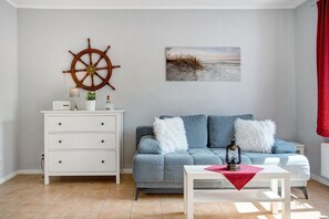 A living room with a sofa, a white side table, a wall-mounted ship's wheel, and a beach-themed painting.