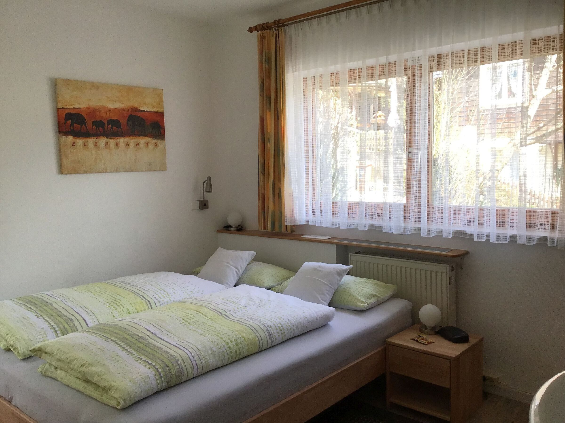 Una cama individual con una manta blanca y verde a rayas, una mesita de noche con lámpara, y una ventana con cortinas translúcidas.