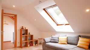 A loft-style room with a sloped ceiling, a built-in wooden bookshelf, a grey sofa with patterned cushions, and a skylight. - Holiday Apartment "Am Eiberg" with Mountain View, Private Terrace & Wi-Fi (Bad Wildbad)