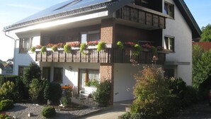 A two-story building with a sloped roof, a balcony with flower boxes, and a paved driveway. - Apartment 'Schelb' with Mountain View, Balcony and Wi-Fi (Sasbachwalden)