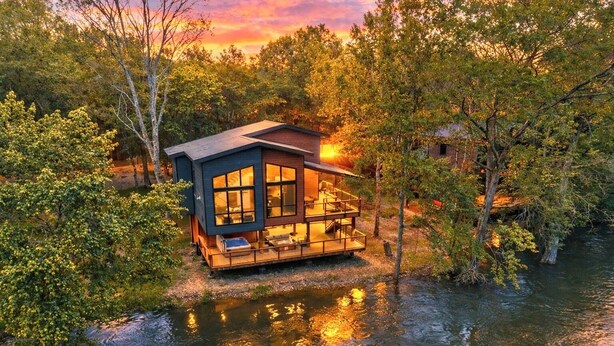 A modern house with a deck overlooking a body of water, surrounded by trees.